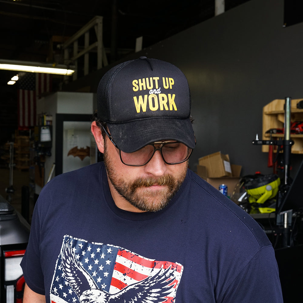 Black flat bill trucker hat with black mesh side panels and "Shut Up and Work" in gold and white lettering across the front.