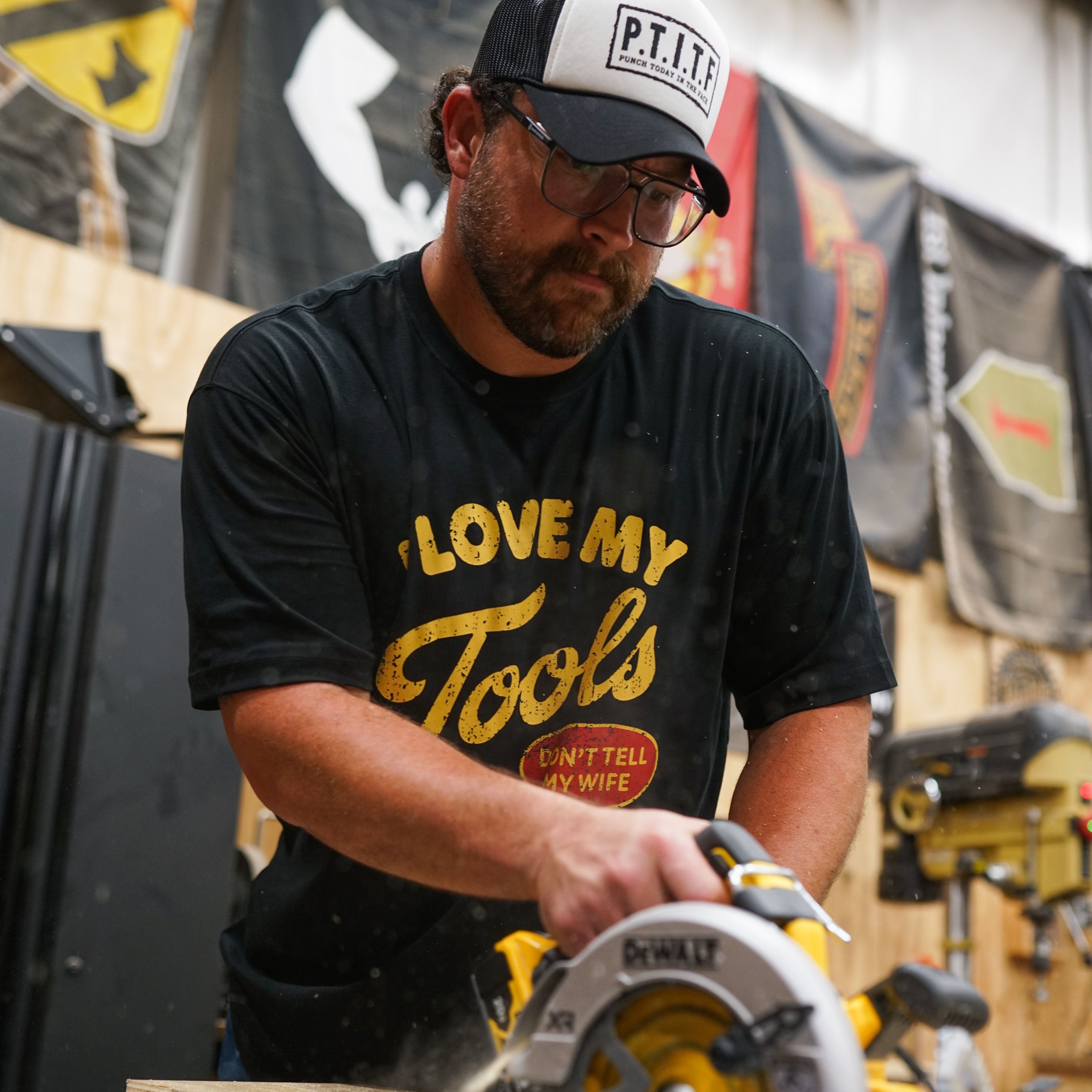 A man wearing a black t-shirt with a yellow graphic that reads "I Love My Tools" in vintage gold lettering and "Don't Tell My Wife" in a red oval badge below.