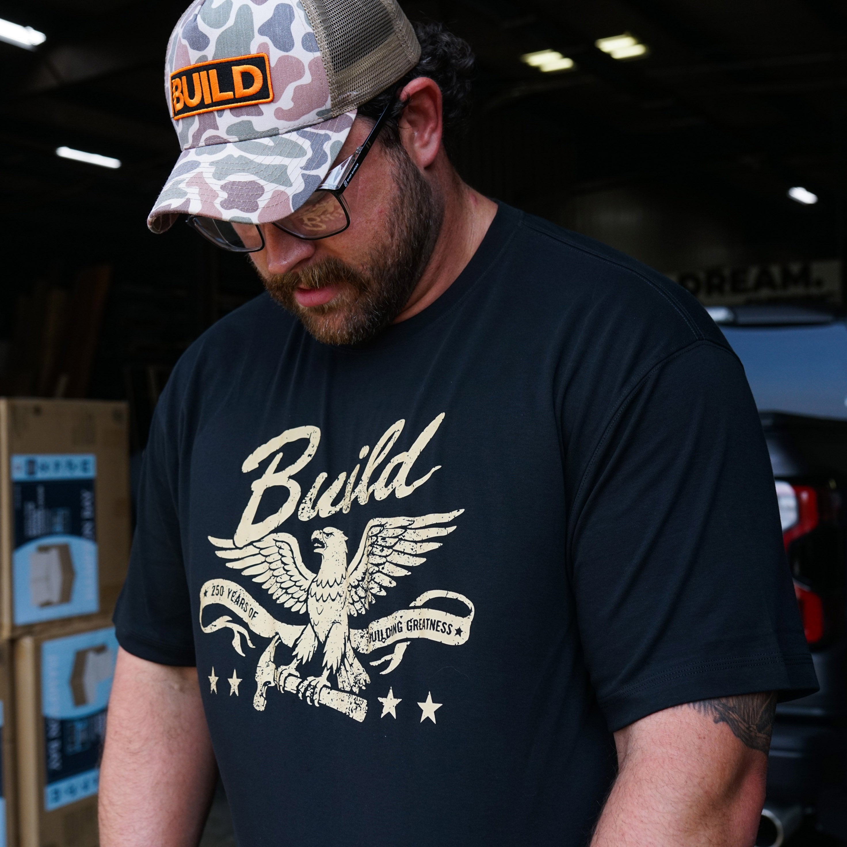 Black t-shirt with a gold eagle perched on a hammer, "Build" in script above, and a banner reading "250 Years of Building Greatness" with four stars below.